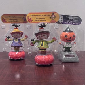 May include: Three solar-powered Halloween decorations: two scarecrows and a pumpkin. The scarecrows have hats, and the pumpkin wears a witch's hat. Each figure is on a red base and has the text "Solar Dancing Scarecrow" or "Solar Dancing Pumpkin".
