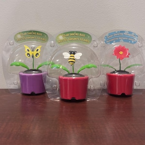 May include: Three solar-powered dancing bug toys in plastic packaging. The toys feature a butterfly, a bee, and a poinsettia flower, each in a small pot. The pots are purple and red. The text on the packaging reads "Solar Dancing Bugs" and "Poinsettia dansant solaire."