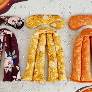 May include: Three sets of fabric-covered heat packs, each with a matching eye mask. The heat packs are shaped like long, narrow pillows with multiple chambers. The eye masks are rectangular and have a floral pattern. The heat packs are in burgundy, yellow, and orange with floral patterns.