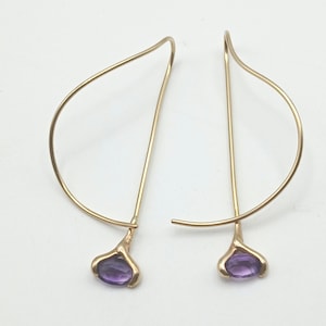 May include: A pair of gold-toned earrings featuring a curved, open-wire hook design. Each earring terminates in a small, prong-set oval purple gemstone at the base, creating a drop-style silhouette with a polished metallic finish.