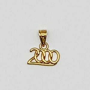 May include: A gold-toned pendant shaped as the number 2000, featuring a stylized font with interconnected loops for the zeros and a triangular base for the two. The piece includes a small, integrated bail at the top for attachment to a chain.