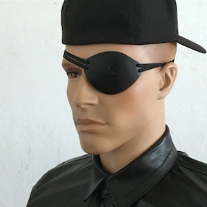 Peut inclure: Un cache-œil en cuir noir avec une sangle réglable. Le cache-œil est porté sur l'œil droit d'une personne portant une casquette de baseball noire.