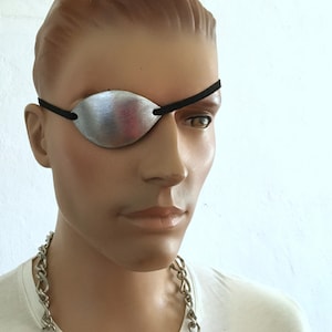 Puede incluir: Un parche de ojo plateado con una correa elástica negra. El parche se lleva sobre el ojo izquierdo.