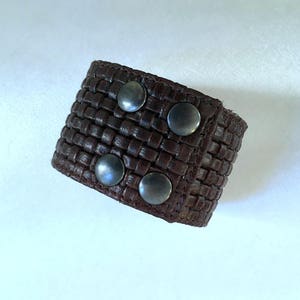 Puede incluir: Un brazalete de cuero marrón con un diseño tejido y cuatro tachuelas de metal plateado.