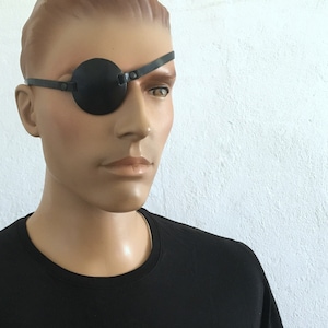 Peut inclure: Un cache-œil en cuir noir avec une sangle qui passe autour de la tête. Le cache-œil est porté sur l'œil droit.