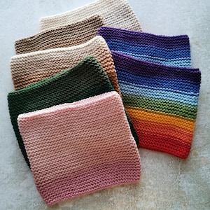 Könnte beinhalten: Eine Sammlung gestrickter quadratischer Tücher in verschiedenen Farben, darunter Creme, Braun, Grün, Rosa und ein Regenbogenverlauf. Die Tücher sind fächerförmig angeordnet und zeigen ihre strukturierte Oberfläche und handgefertigte Optik. Jedes Tuch misst ca. 20 cm x 20 cm.