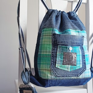 Puede incluir: Una mochila de lona azul con cordón de ajuste, un bolsillo de cuadros verdes y azules y un bolsillo de lona más pequeño con un parche de lentejuelas plateadas. La mochila tiene correas blancas y azules.