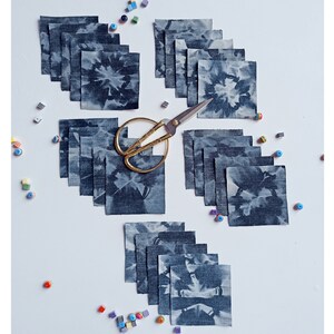 Può includere: Una collezione di 20 quadrati di tessuto denim blu con un motivo a foglie bianche. I quadrati sono disposti in due file da dieci, con un paio di forbici dorate al centro.