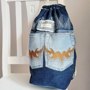 Op de afbeelding: Een blauwe denim rugzak met een trekkoord en twee grote zakken aan de voorkant. De zakken hebben bruine geborduurde ontwerpen en de rugzak heeft een klein label met de tekst "garyusha".