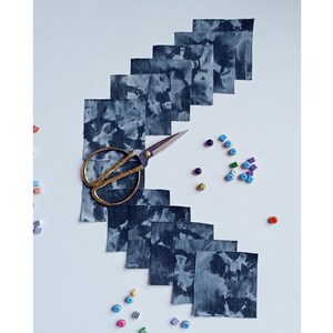 May include: A set of 10 blue fabric squares with a white tie-dye pattern arranged in a staggered pattern on a white surface. A pair of gold scissors is in the center of the squares. There are colorful beads scattered around the squares.