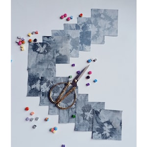 Può includere: Una collezione di quadrati di tessuto blu e bianco con un motivo tie-dye, disposti a semicerchio su una superficie bianca. Un paio di forbici dorate e perline colorate sono sparse intorno ai quadrati di tessuto.