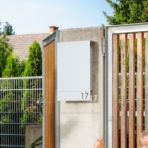 May include: A woman in a black top and jeans stands outside a house, holding a piece of paper. A young girl in a black dress and a pink flower in her hair is playing with a hula hoop in front of a wooden gate.