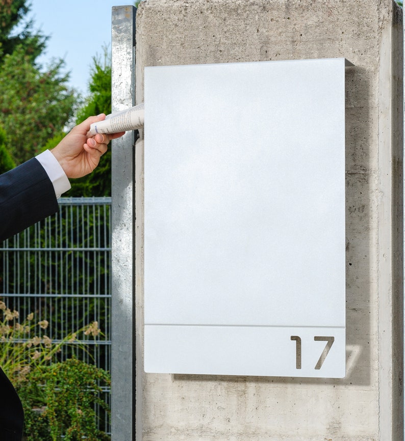 May include: A white metal mailbox with the number 17 cut out of the front. A person is inserting a rolled up newspaper into the mailbox.