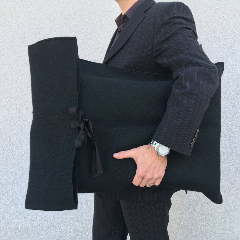 May include: A black fabric cushion with a ribbon tied around it. The cushion is being held by a person wearing a black suit.