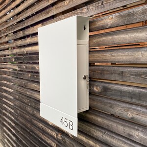 May include: White metal mailbox with a door and a keyhole. The mailbox is mounted on a wooden wall and has the number 45B printed on the front.