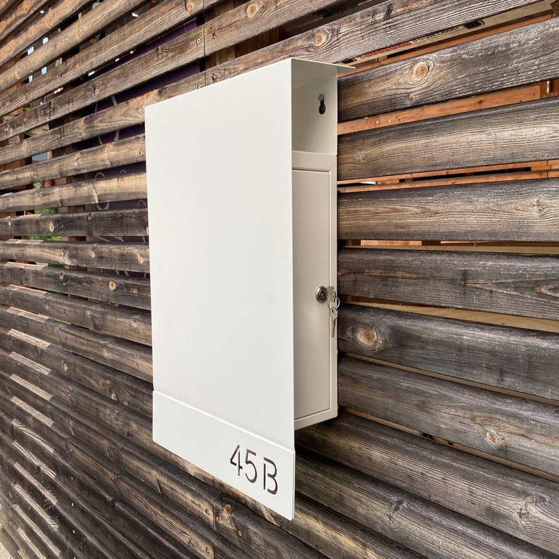May include: White metal mailbox with a door and a keyhole. The mailbox is mounted on a wooden wall and has the number 45B printed on the front.