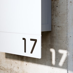 May include: White mailbox with the number 17 cut out of the front. The mailbox is mounted to a concrete wall.