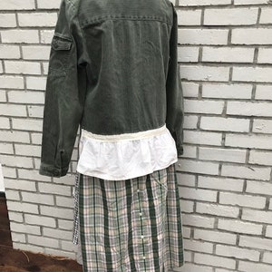 May include: An olive green jacket with a white ruffled hem is paired with a plaid skirt in shades of green, pink, and white. The skirt has a button-down front. The outfit is displayed on a white mannequin.