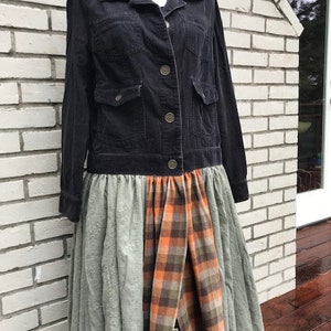 Op de afbeelding: Een zwarte corduroy jas met vier voorzakken en knopen, gecombineerd met een patchwork rok. De rok heeft panelen van saliegroen kant en een ruitpatroon in tinten oranje, bruin en groen.