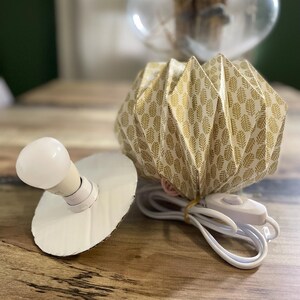 K&ouml;nnte beinhalten: Ein gefalteter Papierlampenschirm mit goldenem Blattmuster, begleitet von einer wei&szlig;en Gl&uuml;hbirne auf einer runden Pappbasis und einem wei&szlig;en Stromkabel mit Schalter. Der Schirm hat ein geometrisches Design in Ziehharmonika-Optik.