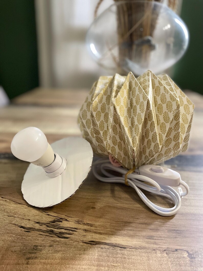 K&ouml;nnte beinhalten: Ein gefalteter Papierlampenschirm mit goldenem Blattmuster, begleitet von einer wei&szlig;en Gl&uuml;hbirne auf einer runden Pappbasis und einem wei&szlig;en Stromkabel mit Schalter. Der Schirm hat ein geometrisches Design in Ziehharmonika-Optik.