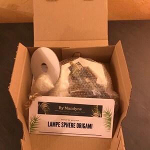 K&ouml;nnte beinhalten: Ein Versandkarton aus Pappe mit einer wei&szlig;en runden Spule, einem kleinen eingewickelten B&uuml;ndel und einem in Luftpolsterfolie verpackten Gegenstand. Eine wei&szlig;e Karte mit schwarzem Text zeigt By Mandyne, Notice de Montage, LAMPE SPHERE ORIGAMI.