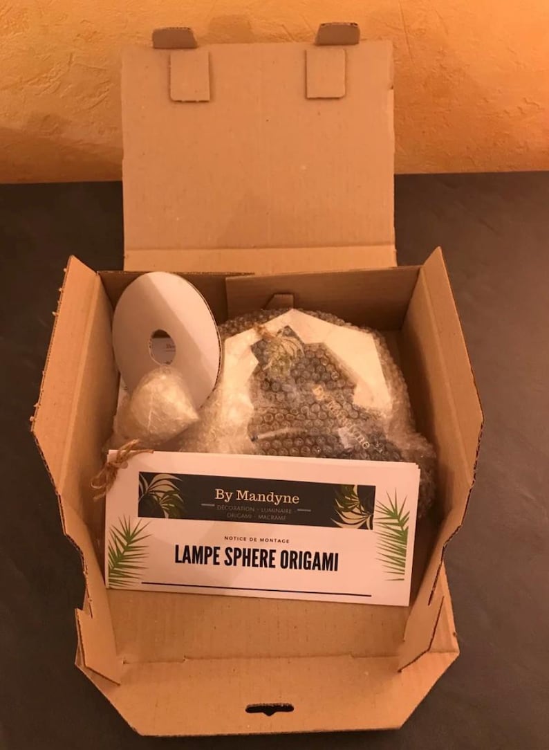 K&ouml;nnte beinhalten: Ein Versandkarton aus Pappe mit einer wei&szlig;en runden Spule, einem kleinen eingewickelten B&uuml;ndel und einem in Luftpolsterfolie verpackten Gegenstand. Eine wei&szlig;e Karte mit schwarzem Text zeigt By Mandyne, Notice de Montage, LAMPE SPHERE ORIGAMI.