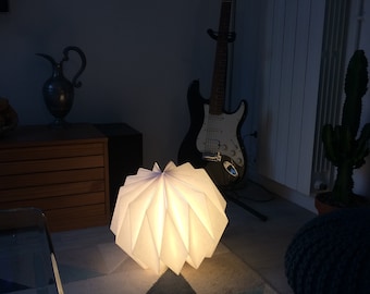 Könnte beinhalten: Eine weiße Lampe im Origami-Stil mit einem Sternmuster beleuchtet einen Raum mit einem gemusterten Teppich, einer Gitarre und einem Topf-Kaktus.