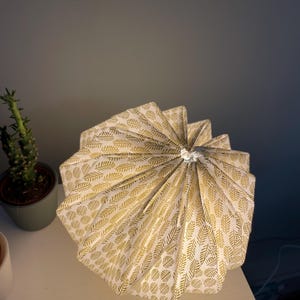 K&ouml;nnte beinhalten: Ein plissierter Lampenschirm mit wei&szlig;em Hintergrund und einem sich wiederholenden goldenen Blattmuster. Der Schirm hat ein kreisf&ouml;rmiges, gefaltetes Design, das in einem Mittelpunkt zusammenl&auml;uft und eine texturierte F&auml;cheroptik erzeugt.