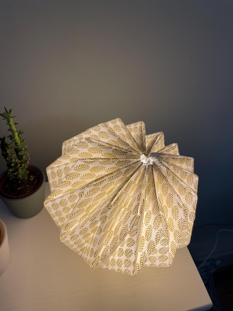 K&ouml;nnte beinhalten: Ein plissierter Lampenschirm mit wei&szlig;em Hintergrund und einem sich wiederholenden goldenen Blattmuster. Der Schirm hat ein kreisf&ouml;rmiges, gefaltetes Design, das in einem Mittelpunkt zusammenl&auml;uft und eine texturierte F&auml;cheroptik erzeugt.