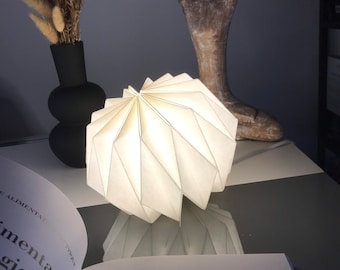 Könnte beinhalten: Eine weiße Papierlampe mit gefaltetem Design, die leuchtet und einen warmen Schein erzeugt. Die Lampe steht auf einer dunklen Oberfläche neben einer Holzskulptur einer Person in einer Yogapose.