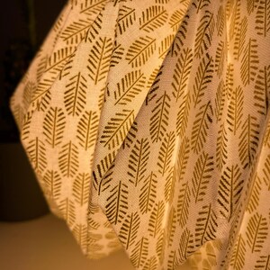 K&ouml;nnte beinhalten: Ein plissierter Lampenschirm aus cremefarbenem Stoff mit einem sich wiederholenden Muster aus dunkelgoldenen Farnblattmotiven. Das Material hat eine sichtbare Webstruktur und die Struktur wird von innen beleuchtet, was einen warmen Schein wirft.