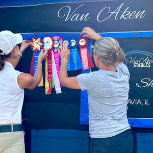 May include: Two people are standing in front of a blue and black wall with the text "Van Aken Stables" and "Shiloh, IL" on it. They are holding up ribbons with the words "Champion", "Reserve Champion", "First Place", "Second Place", and "Third Place" on them.