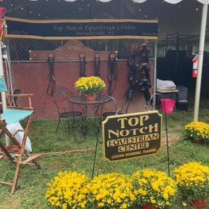 May include: A black sign with gold lettering reads "Top Notch Equestrian Centre" with the tagline "Lessons-Boarding-Training". The sign is in front of a white tent with a banner that reads "Top Notch Equestrian Centre, Stewartsville, NJ". There are yellow mums in pots in front of the sign.