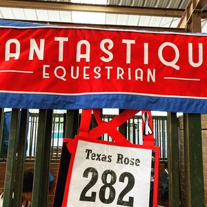 May include: A red banner with white text that reads "Fantastique Equestrian" hanging above a stall with a white sign that reads "Texas Rose 282".