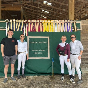 May include: Four people stand in front of a green wall with a sign that reads "Limerick Lane Farm, Maureen A. Fagan, Hilliard, Ohio". The wall is decorated with a row of colorful ribbons. The people are wearing casual clothing.