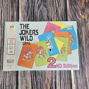 Może przedstawiać: Zabytkowe pudełko gry "The Joker's Wild Game", 2. wydanie, z kolorowymi kartami do gry z postaciami z kreskówek. Pudełko ma logo Milton Bradley Company i tekst "Ages 10 to Adult".