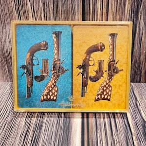 Puede incluir: Dos cartas de juego vintage que presentan un par de pistolas antiguas con diseños intrincados. Las cartas están en un esquema de color azul y dorado con el texto "Hallmark" impreso en la parte inferior.