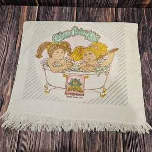 May include: A white bath towel with a fringe border features a Cabbage Patch Kids illustration of two dolls taking a bath in a white bathtub with green and pink accents. The text "Cabbage Patch Kids" is printed in a green and pink font above the dolls.