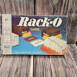May include: A vintage Rack-O board game box with the game title in large white letters. The box features illustrations of game components, including cards and racks, on a blue background. The box is on a wooden surface.