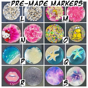 May include: Nine round resin coasters with various designs. The coasters feature a variety of themes, including seashells, cartoon characters, and abstract patterns. The coasters are labeled with letters L through S.