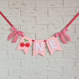 Peut inclure: Une guirlande rose avec le mot "ONE" en lettres blanches, ornée de cerises rouges et de nœuds. La guirlande est suspendue par un ruban rouge, sur un mur de briques blanches.