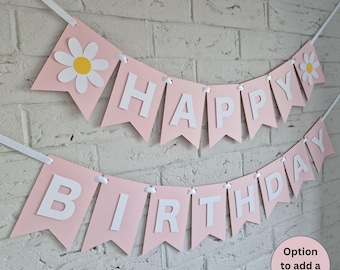 Op de afbeelding: Een roze banner met de woorden "HAPPY BIRTHDAY" in witte letters. De banner is versierd met witte madeliefjes en geregen met wit lint. De tekst "Option to add a name!" staat in een roze cirkel.