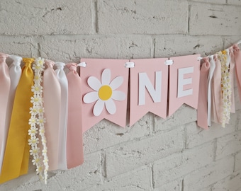 Könnte beinhalten: Ein dekoratives Banner mit hellrosa Wimpeln und weißen Buchstaben, die das Wort ONE bilden, sowie einem weißen und gelben Gänseblümchen-Ausschnitt auf dem ersten Wimpel. Das Banner ist mit gelben, weißen und rosa Bändern sowie Gänseblümchenborte verziert.