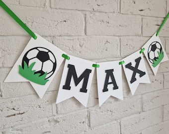 May include: A white banner with the name "MAX" in black letters, interspersed with soccer ball and grass cutouts. The banner is strung with green ribbon and hangs against a white brick wall. A festive decoration.