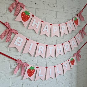 K&ouml;nnte beinhalten: Ein rosa Geburtstagsbanner mit wei&szlig;en Buchstaben, die "HAPPY BIRTHDAY LILY" schreiben. Das Banner ist mit roten Erdbeeren und rot-wei&szlig; karierten Schleifen verziert. Es h&auml;ngt vor einer wei&szlig;en Backsteinmauer.