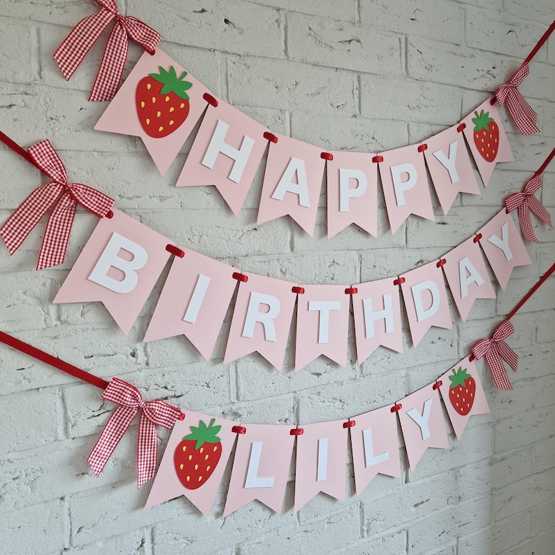 K&ouml;nnte beinhalten: Ein rosa Geburtstagsbanner mit wei&szlig;en Buchstaben, die "HAPPY BIRTHDAY LILY" schreiben. Das Banner ist mit roten Erdbeeren und rot-wei&szlig; karierten Schleifen verziert. Es h&auml;ngt vor einer wei&szlig;en Backsteinmauer.