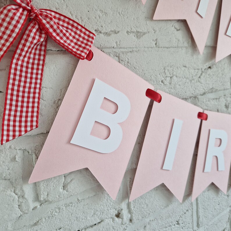 K&ouml;nnte beinhalten: Rosa Banner mit wei&szlig;en Buchstaben, die "BIRTHDAY" buchstabieren. Das Banner ist mit einer rot-wei&szlig; karierten Schleife verziert. Es h&auml;ngt vor einer wei&szlig;en Backsteinmauer.