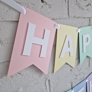 Peut inclure: Banni&egrave;re en papier rose pastel, jaune, vert et bleu avec des lettres blanches qui &eacute;pellent "HAPPY".