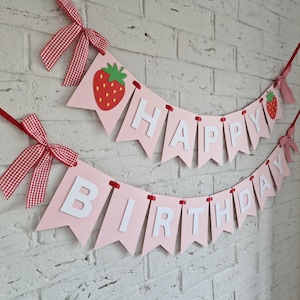 K&ouml;nnte beinhalten: Rosa Banner mit den Worten "HAPPY BIRTHDAY" in wei&szlig;en Buchstaben. Das Banner ist mit roten Erdbeeren und rot-wei&szlig; karierten Schleifen verziert. Das Banner h&auml;ngt an einer wei&szlig;en Backsteinmauer.